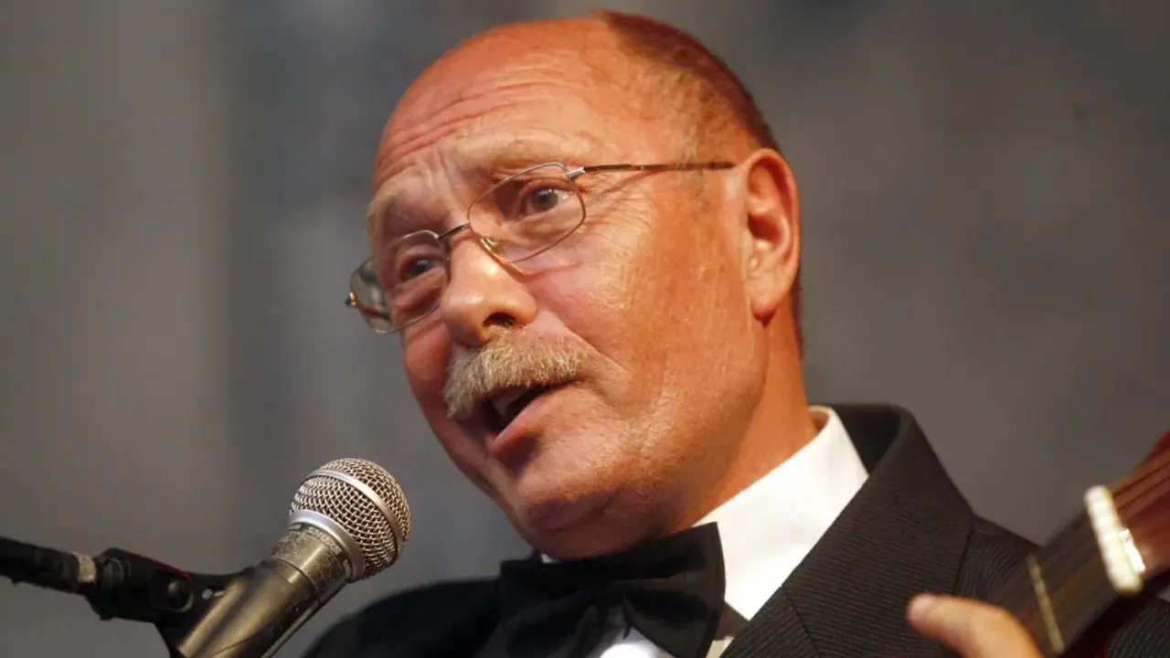 Alosza Awdiejew w czarnym garniturze i muszce śpiewa do mikrofonu, grając na gitarze.