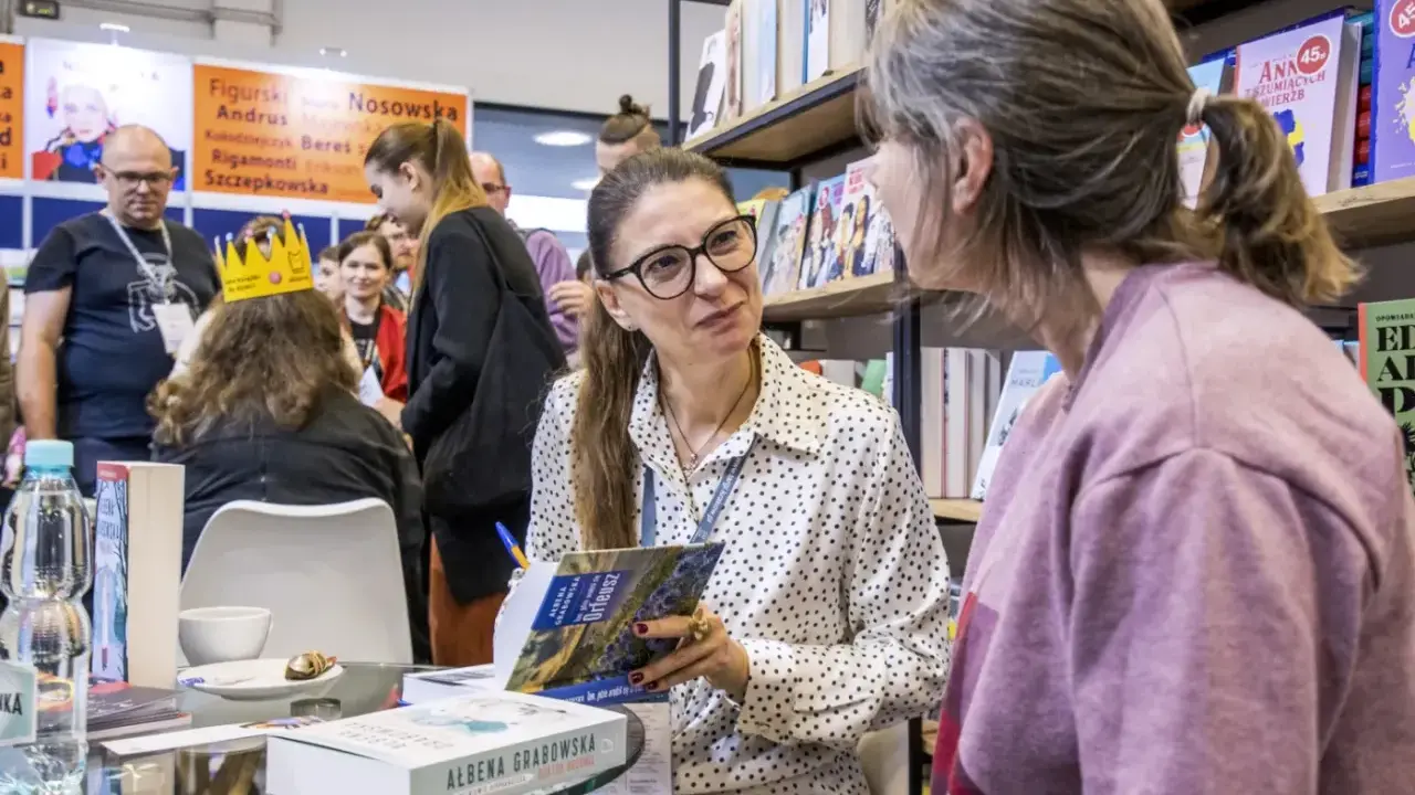 Ałbena Grabowska rozmawia z czytelniczką przy stoisku z książkami. W tle widać innych ludzi i półki z publikacjami.