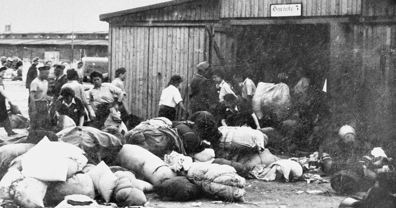 Ludzie z bagażami przed barakiem w Auschwitz. W tle widać innych ludzi i budynki.