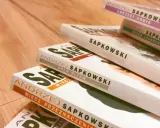 Książki Andrzeja Sapkowskiego ułożone w stos. Widoczne tytuły: "Miecz Przeznaczenia", "Krew Elfów", "Czas Pogardy", "Wieża Jaskółki", "Chrzest Ognia". Idealne do poznania kolejności czytania o wiedźminie.