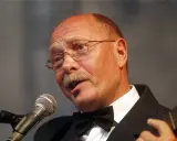 Alosza Awdiejew w czarnym garniturze i muszce śpiewa do mikrofonu, grając na gitarze.