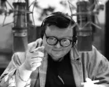 Wojciech Mann w młodości, w słuchawkach i okularach, wskazuje palcem, gotowy do nagrania.