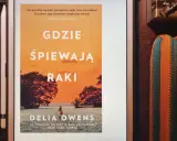 Okładka książki "Gdzie śpiewają raki" z Delia Owens. Na pomarańczowym tle zachodzącego słońca, widać sylwetki drzew i kajakarza na wodzie.