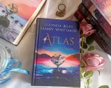 Książka "Atlas. Historia pa Salta" z serii "Siedem sióstr" Lucindy Riley i Harry'ego Whittakera, obok innych tomów cyklu i róż.