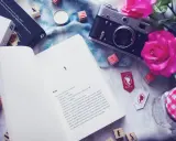 Otwarta książka z tekstem o kolorach życia, obok aparat, róże i kostki z literami. Czytasz o Maxton Hall?