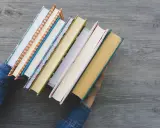 Ręce w dżinsowej kurtce trzymają stos kolorowych książek. Zastanawiasz się, jaką książkę warto przeczytać?