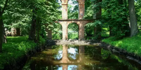 Kamienne łuki i ceglane ruiny tworzą malowniczy motyw arkadii nad spokojną wodą, otoczone bujną zielenią.