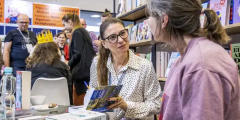 Ałbena Grabowska rozmawia z czytelniczką przy stoisku z książkami. W tle widać innych ludzi i półki z publikacjami.