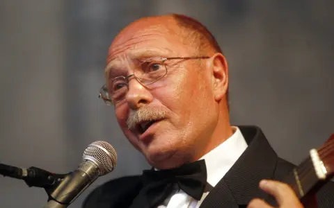 Alosza Awdiejew w czarnym garniturze i muszce śpiewa do mikrofonu, grając na gitarze.