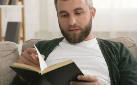 Mężczyzna z brodą czyta książkę, przeglądając strony. Na półce w tle widać **książki chronologicznie**.