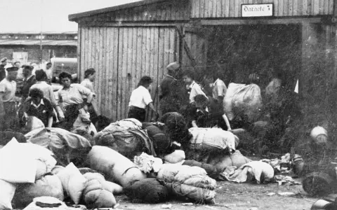 Ludzie z bagażami przed barakiem w Auschwitz. W tle widać innych ludzi i budynki.
