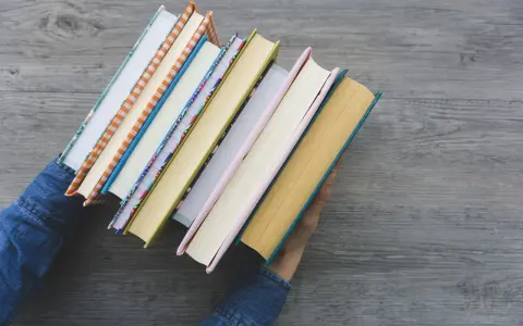 Ręce w dżinsowej kurtce trzymają stos kolorowych książek. Zastanawiasz się, jaką książkę warto przeczytać?