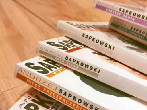 Książki Andrzeja Sapkowskiego ułożone w stos. Widoczne tytuły: "Miecz Przeznaczenia", "Krew Elfów", "Czas Pogardy", "Wieża Jaskółki", "Chrzest Ognia". Idealne do poznania kolejności czytania o wiedźminie.
