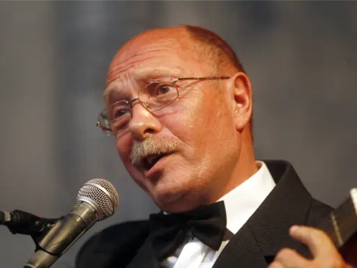 Alosza Awdiejew w czarnym garniturze i muszce śpiewa do mikrofonu, grając na gitarze.