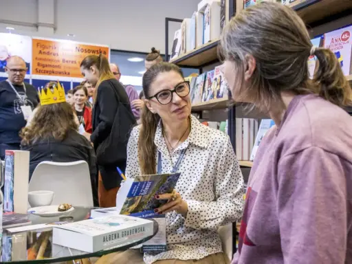 Ałbena Grabowska rozmawia z czytelniczką przy stoisku z książkami. W tle widać innych ludzi i półki z publikacjami.