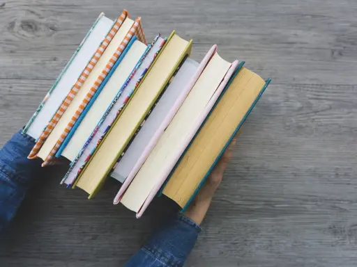 Ręce w dżinsowej kurtce trzymają stos kolorowych książek. Zastanawiasz się, jaką książkę warto przeczytać?