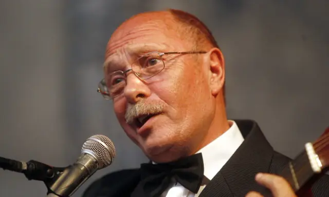 Alosza Awdiejew w czarnym garniturze i muszce śpiewa do mikrofonu, grając na gitarze.