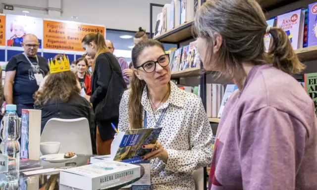 Ałbena Grabowska rozmawia z czytelniczką przy stoisku z książkami. W tle widać innych ludzi i półki z publikacjami.