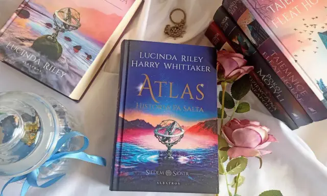Książka "Atlas. Historia pa Salta" z serii "Siedem sióstr" Lucindy Riley i Harry'ego Whittakera, obok innych tomów cyklu i róż.