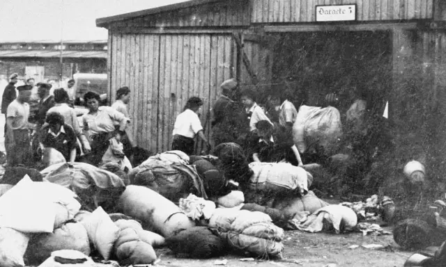 Ludzie z bagażami przed barakiem w Auschwitz. W tle widać innych ludzi i budynki.