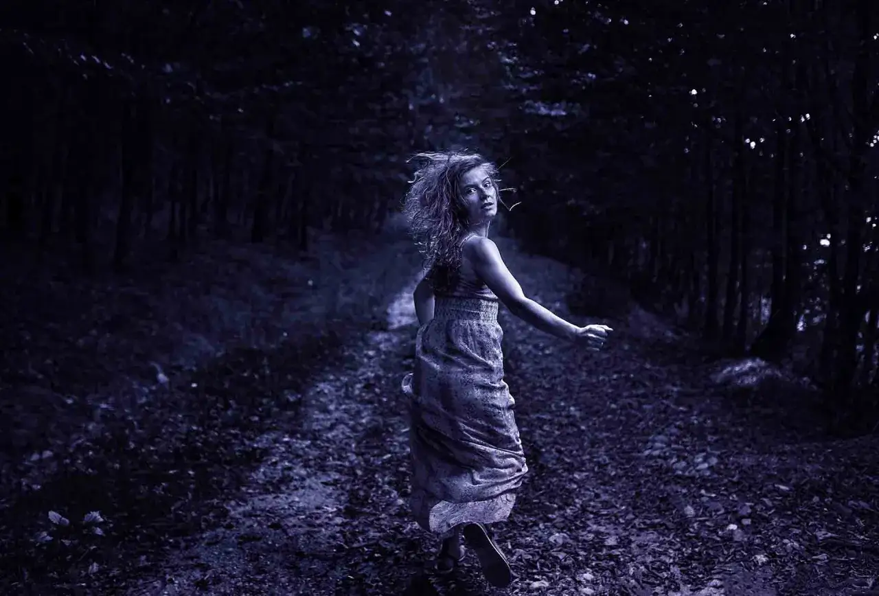 Dziewczyna w zwiewnej sukience biegnie przez mroczny las. Jej spojrzenie pełne jest strachu, jakby uciekała przed czymś niewidzialnym. Scena jak z mrocznego thrillera psychologicznego.