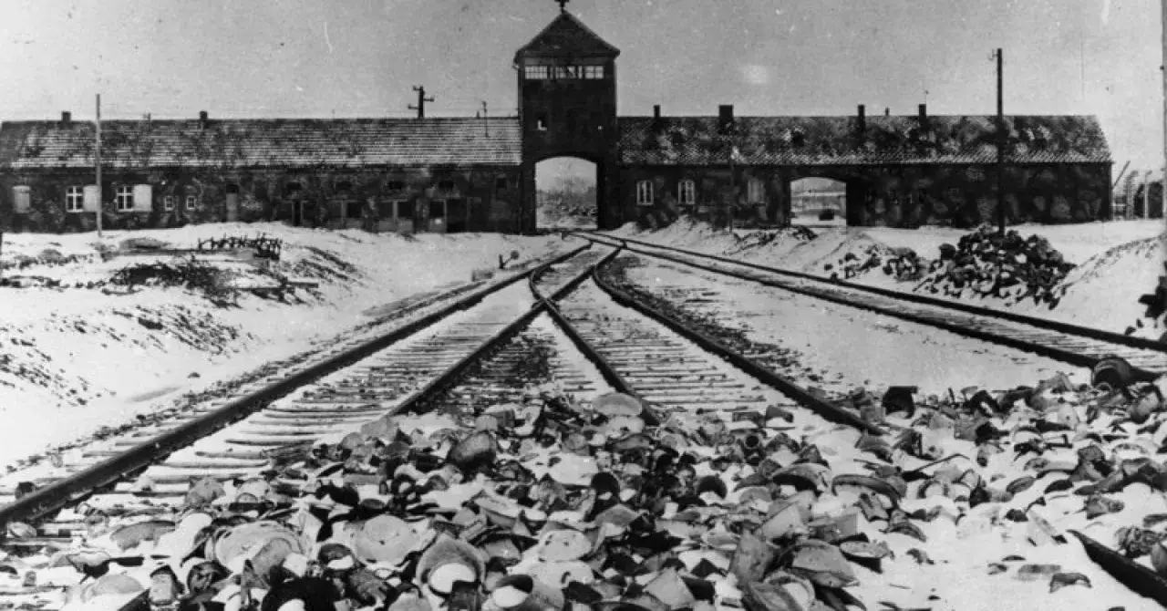 Tory kolejowe prowadzące do bramy obozu Auschwitz, zasypane śniegiem i gruzem. W tle budynek z wieżą.