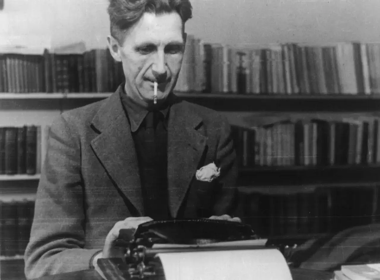 Eric Arthur Blair, znany jako George Orwell, pali papierosa przy maszynie do pisania, otoczony regałami pełnymi książek.