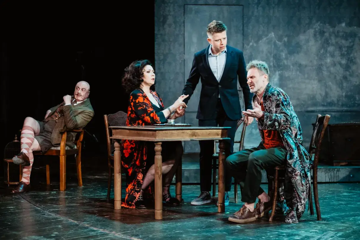 Na scenie cztery postacie. Mężczyzna w garniturze stoi nad kobietą przy stole. Obok siedzi mężczyzna w barwnej szacie. Z boku siedzi starszy pan. Tango streszczenie.